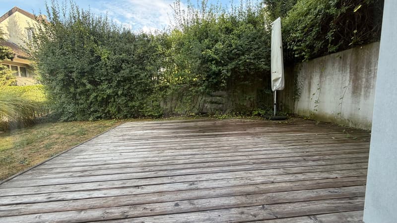Wooden deck area surrounded by greenery and a concrete wall, with an umbrella stand visible.