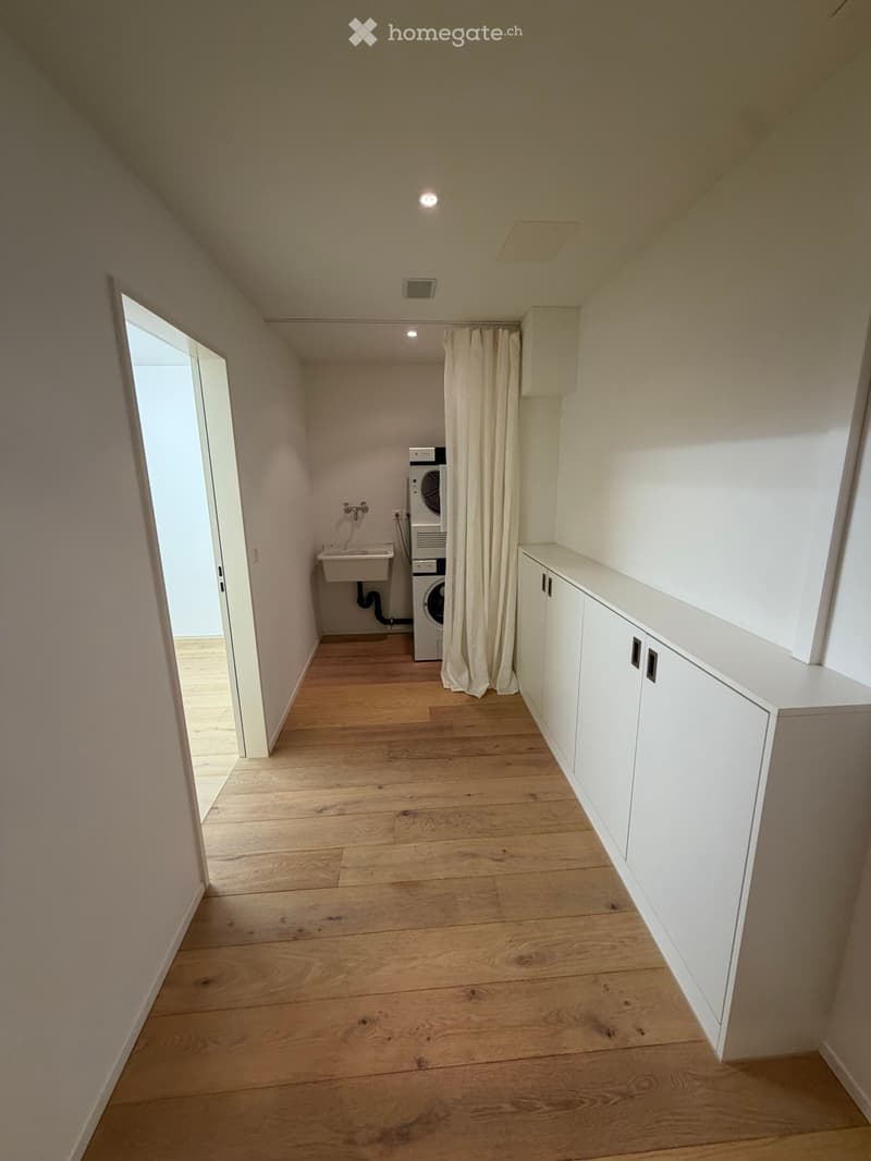 Empty hallway with wooden flooring, white walls, and a small sink area with a washing machine. Features built-in white cabinets and a curtain separating the washing area.