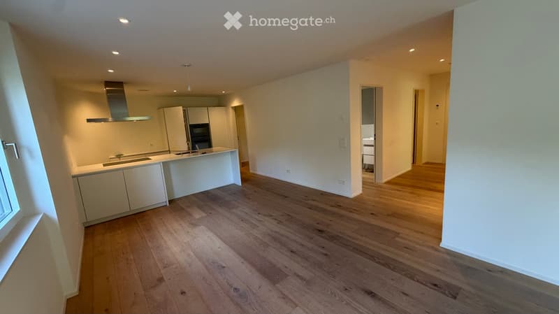 Open-plan kitchen and living area with light wooden flooring, white cabinetry, and a stainless steel range hood. The space is empty, with no furniture or appliances installed.