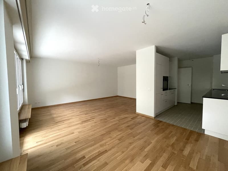 Empty room with wooden flooring, white walls, and a small kitchen area with cabinets and appliances visible in the background.