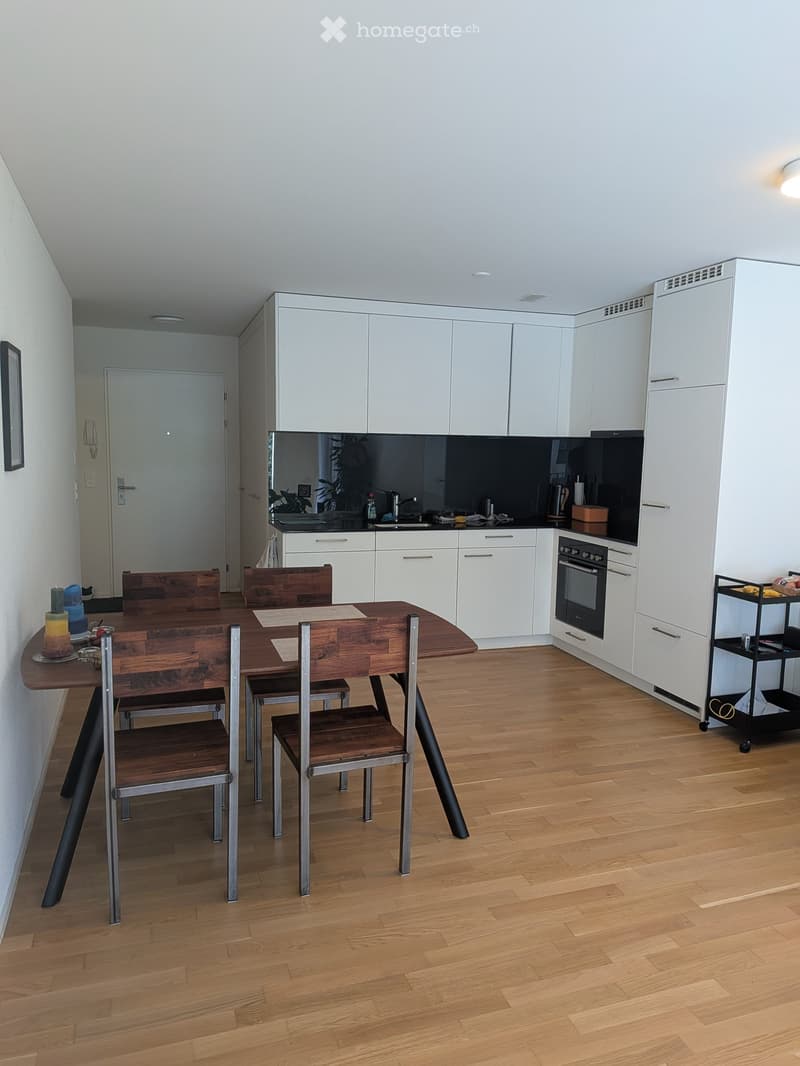 Modern kitchen with white cabinets, dark countertops, wooden dining table with four chairs, and a small cart with items.