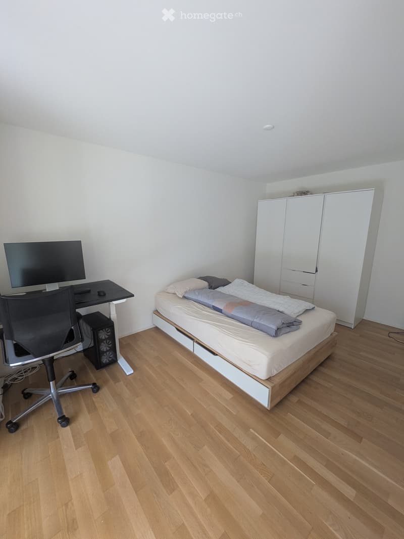 A small, furnished bedroom with a wooden floor, a single bed with pillows and blankets, a desk with a chair and computer screen, and a white wardrobe.