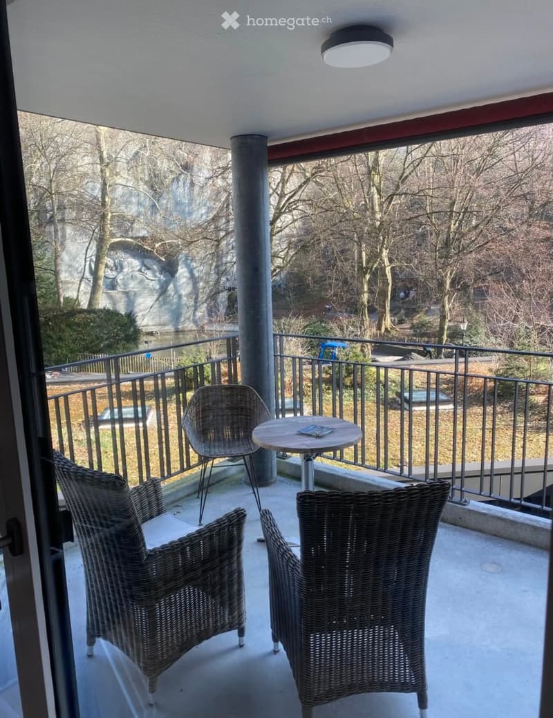 Balcony with a round table and two wicker chairs, overlooking a park with trees and a river.