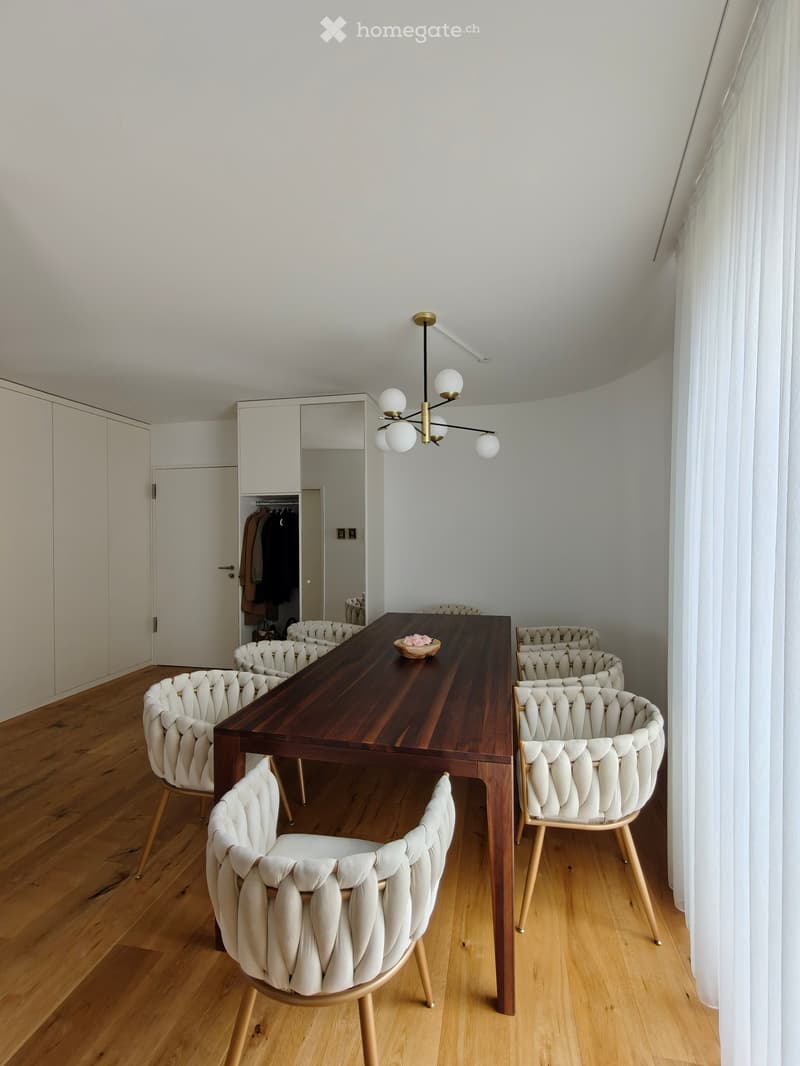 Modern dining area with a long wooden table, six white upholstered chairs, a chandelier, and a large mirror on the wall.