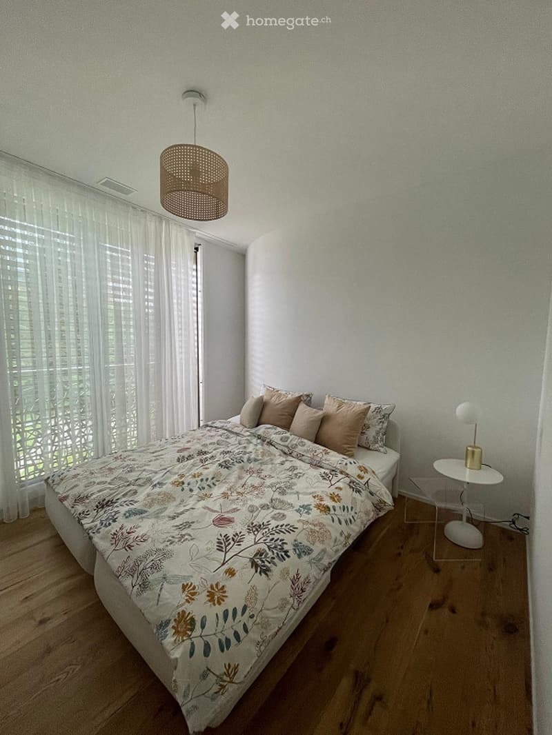 A modern bedroom with a large bed, floral duvet, wooden floor, white walls, and a pendant light hanging from the ceiling. A small nightstand with a table lamp is visible next to the bed, and sheer white curtains cover the window.