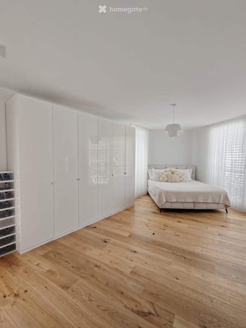 Modern bedroom with light wooden flooring, a large bed with pillows, a tall white wardrobe, a small storage unit, and a hanging pendant light.