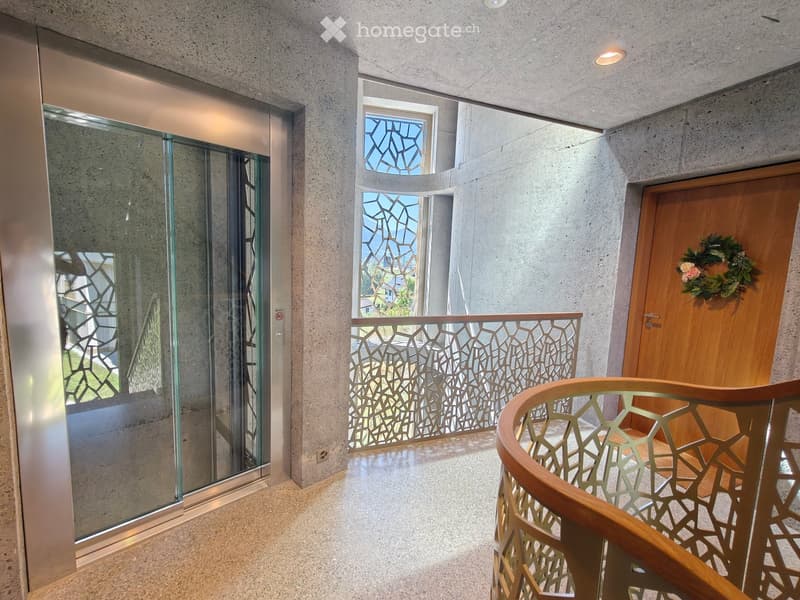 Modern hallway with concrete walls, glass elevator, decorative railing, and a wooden door with a floral wreath.