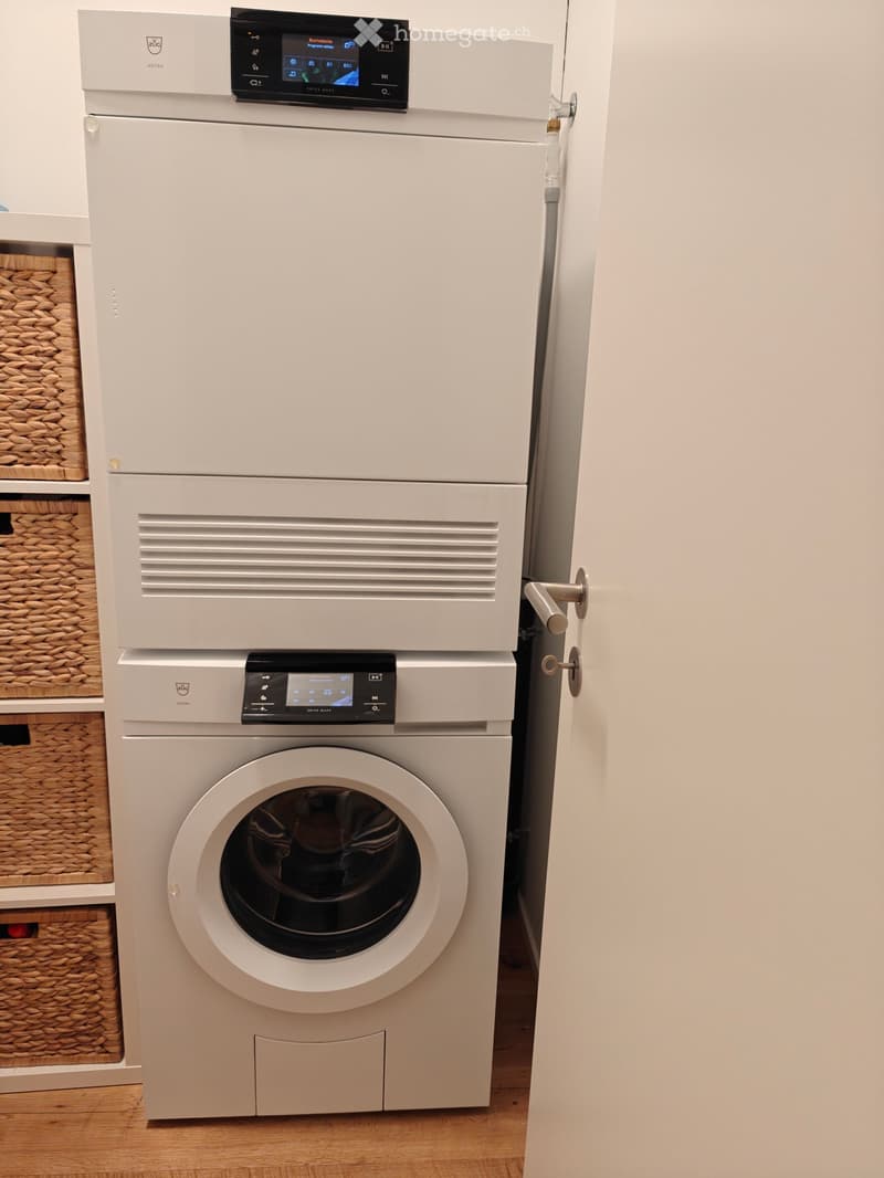 Modern bathroom with a white washing machine and dryer unit, featuring a control panel on top and a wicker basket on the left side.