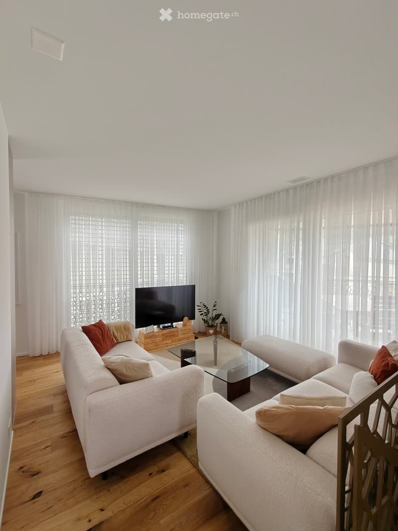 Modern living room with white walls, wooden flooring, large windows with white curtains, a white sectional sofa with red and beige cushions, a glass coffee table, a television on a wooden stand, and a small potted plant.