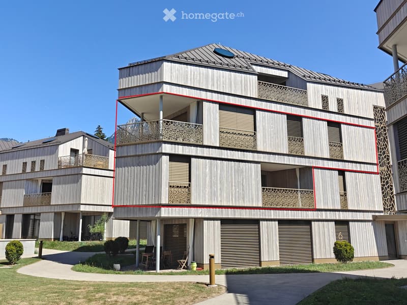 Modern multi-story house with wooden exterior, balconies, and contemporary design. Features include large windows, decorative railings, and a flat roof with solar panels.