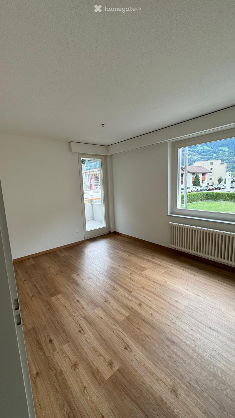 Empty room with wooden flooring, white walls, a radiator, and large windows offering a view of the outdoors.