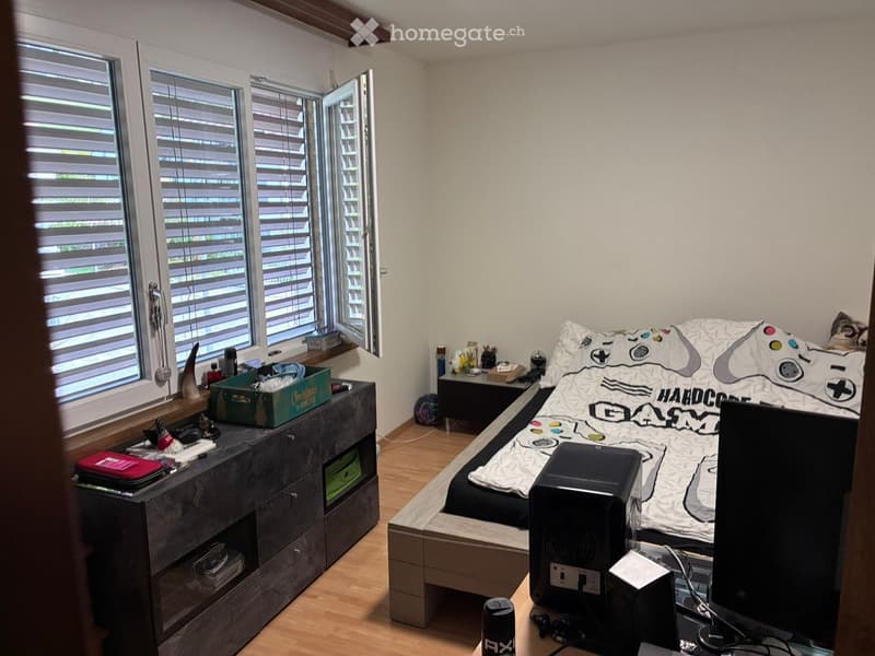 A modern bedroom with a gaming-themed bedspread, a large window with blinds, a wooden floor, a nightstand, and a drawer unit with various items on top.