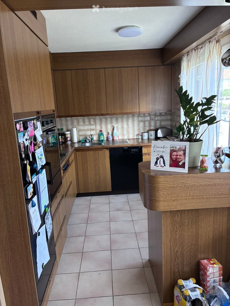 Well-lit kitchen with wooden cabinets, stainless steel appliances, and a central island. Visible items include a refrigerator, oven, dishwasher, sink, toaster, and a framed photo on the counter.