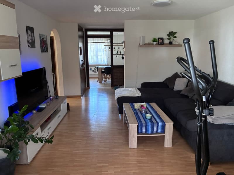 Modern living room with wooden flooring, a dark grey sectional sofa, a light wooden coffee table with a blue and white runner, a TV stand with a flat-screen TV, and a potted plant. An exercise bike is positioned near the sofa.