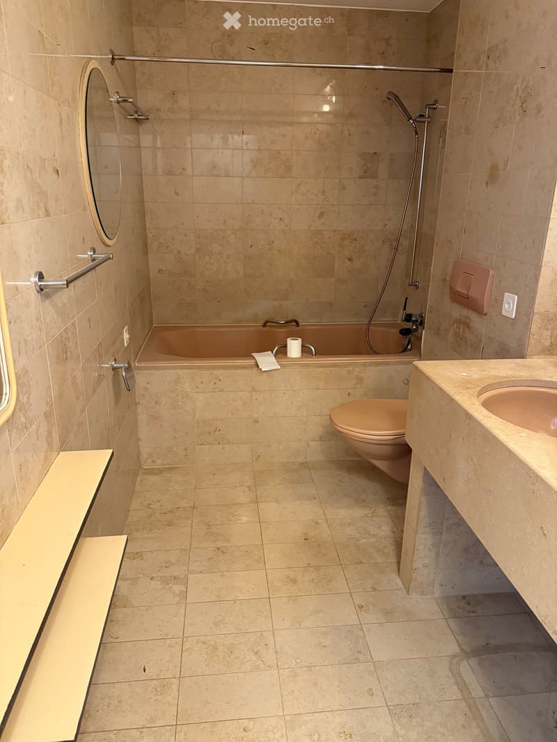 Modern bathroom with beige tiles, a bathtub, toilet, sink, and a towel rack. Features a handheld showerhead and a built-in shelf.