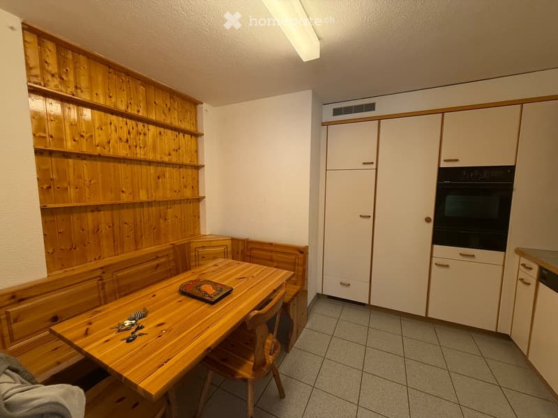 A small kitchen with wooden paneling on one wall, a wooden dining table with a bench seating, and white cabinets with an integrated oven.
