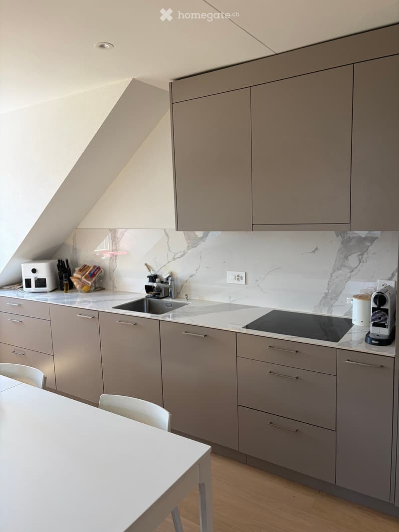 Modern kitchen with grey cabinets, marble backsplash, and stainless steel appliances. Features a sink, coffee machine, and a small dining area with a table and chairs.