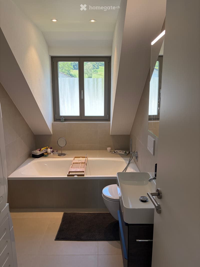 Modern bathroom with a large window, bathtub, sink, toilet, and a small black bath mat on the floor.