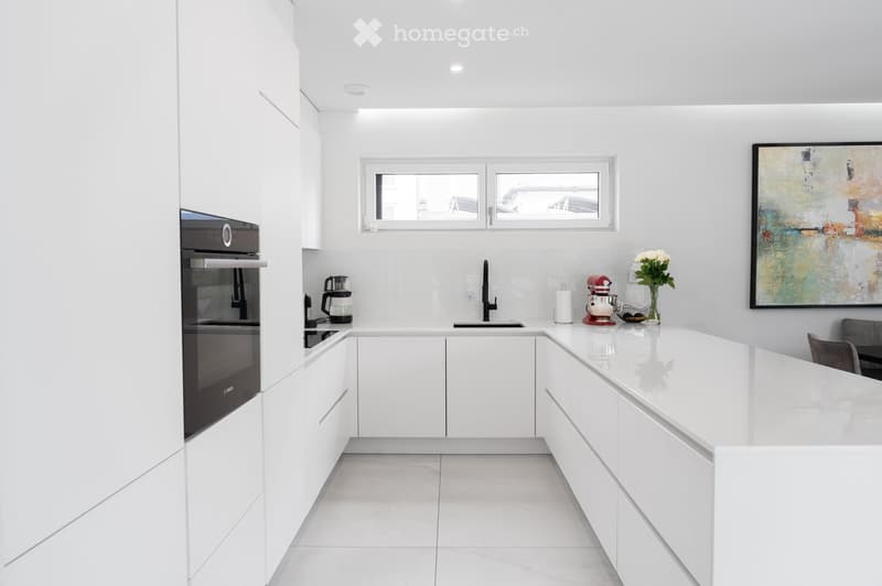Modern white kitchen with sleek cabinetry, a built-in oven, a central island, and a vase with flowers on the counter.
