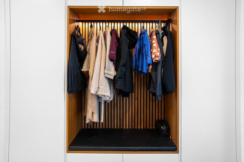 A modern, built-in wardrobe with wooden frame and black bottom shelf, containing various hanging clothes and a small bag on the shelf.