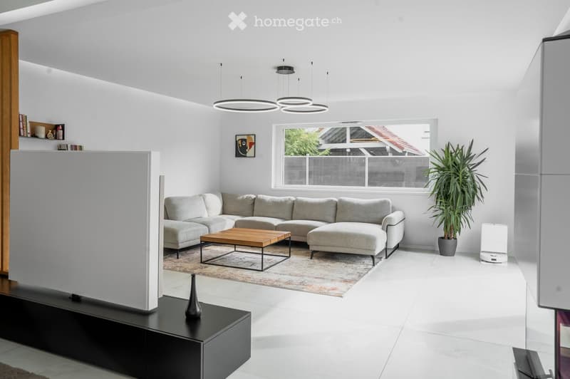 Modern living room with white walls, grey sectional sofa, wooden coffee table, ottoman, floor lamp, potted plant, and a sleek black TV stand.