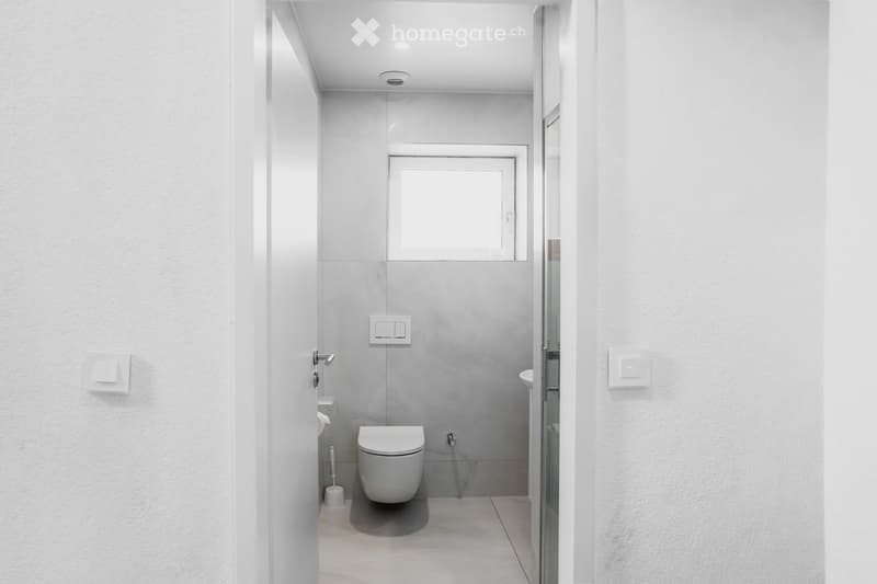 Modern, minimalist bathroom with white walls, grey tiles, a toilet, a sink, and a small window above the toilet.