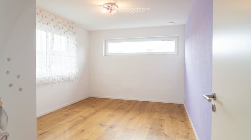 Empty room with wooden flooring, white walls, and two windows. One window has a decorative curtain with butterfly patterns, and the other is a smaller window. The room features a light fixture on the ceiling and a door with a silver handle on the right si