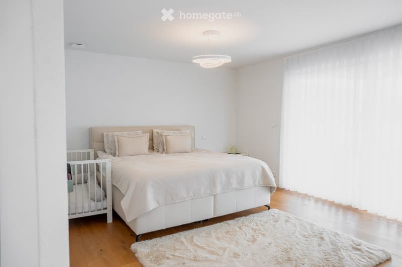Bright bedroom with a large bed, nightstand, and a small baby crib. The room features wooden flooring, white walls, and a modern pendant light.