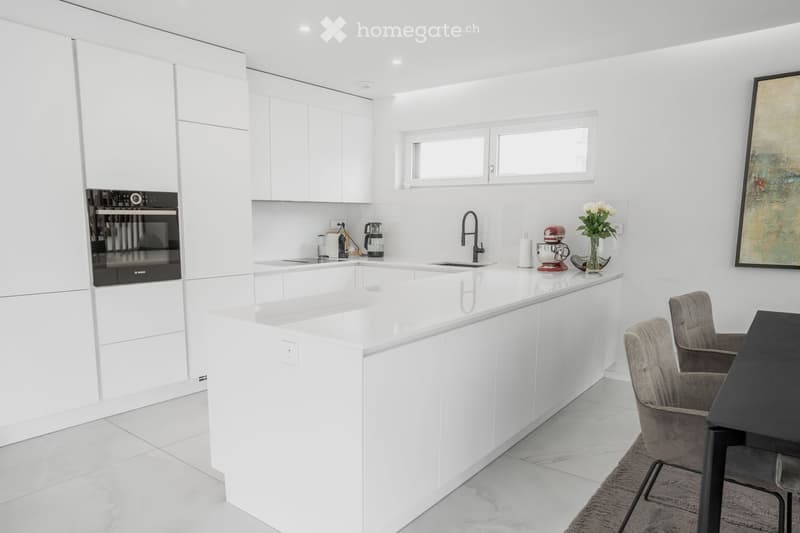 Modern white kitchen with sleek cabinets, central island, built-in oven, sink, and various appliances. Includes a dining area with chairs and a table.