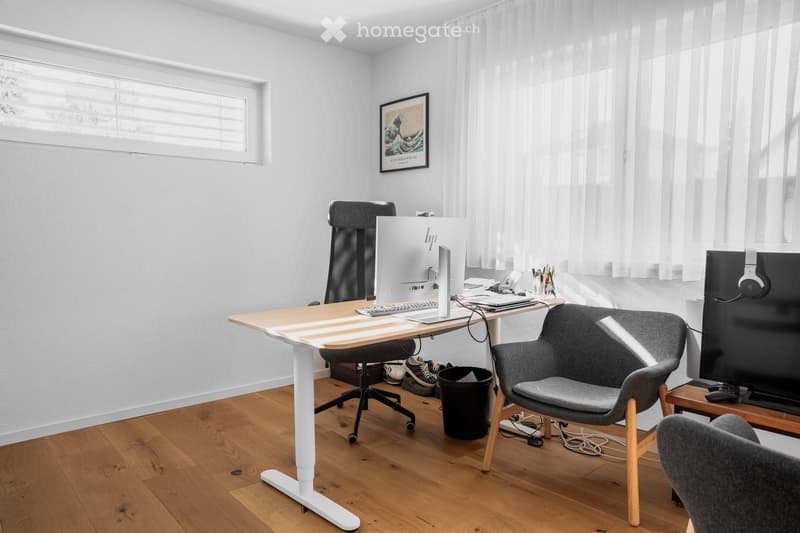 Modern, well-lit room with a standing desk, ergonomic chair, laptop, keyboard, headphones, and a comfortable seating area with a TV and additional chair.