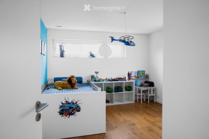 A child's bedroom with a bed, a large stuffed lion toy, shelves filled with books and toys, a small table and chairs, and a globe. The room features wooden flooring and white walls with a blue accent wall.