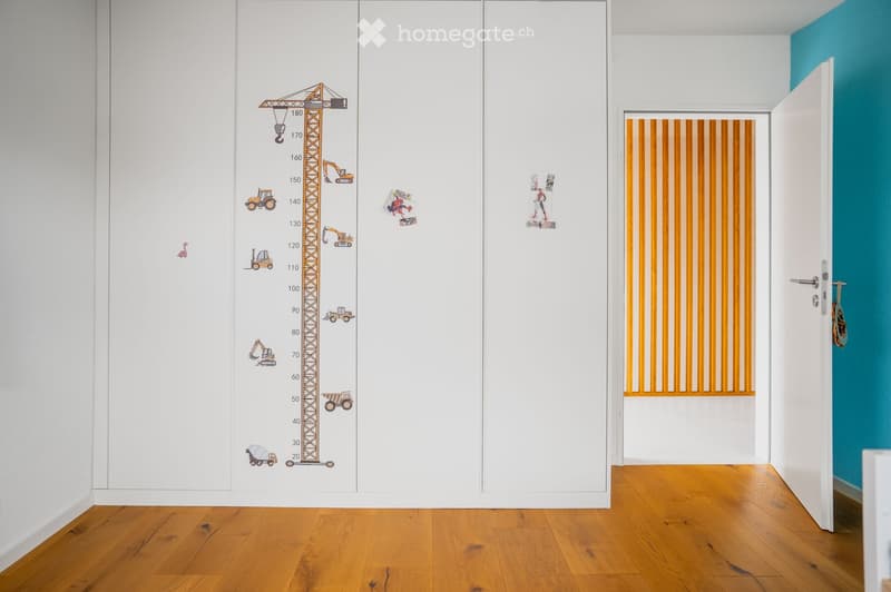 A narrow hallway with white walls, wooden flooring, and a height chart mural featuring construction vehicles. A door leads to another room with orange vertical blinds.