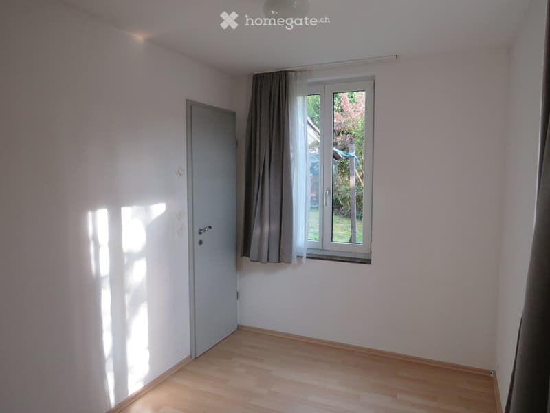 Empty room with light-colored walls, wooden flooring, a door, and a window with curtains. Sunlight streams in from the window.