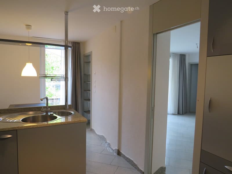 Modern kitchen with a double sink, sleek faucet, built-in cabinets, and a window with curtains. The space is bright and open, with a view of a balcony area.