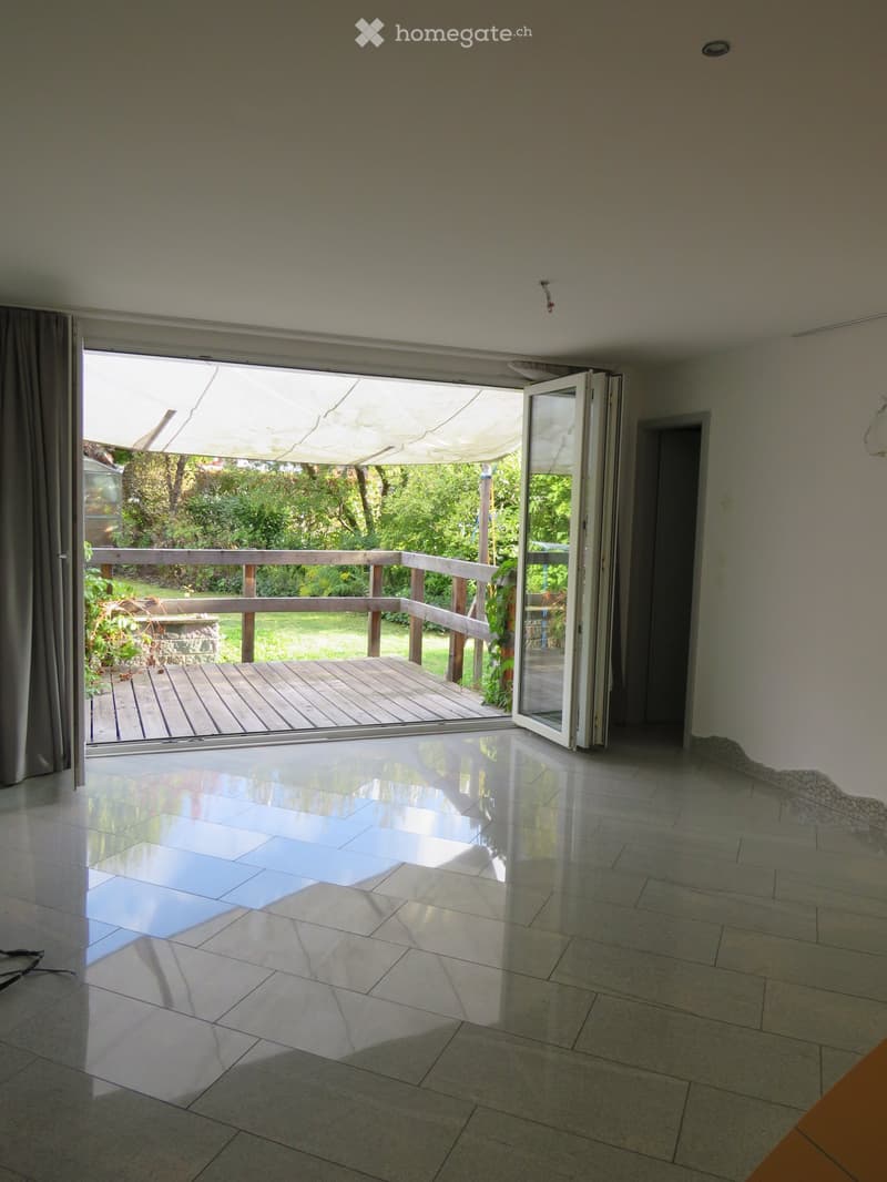 Empty room with large sliding glass doors leading to a wooden deck and garden, tiled floor, and white walls.