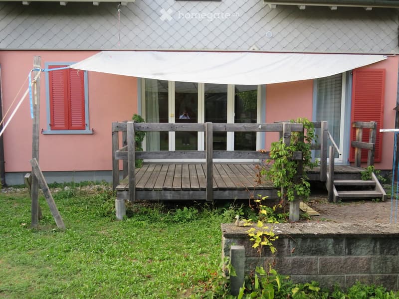 Wooden deck attached to a pink house with red shutters, featuring a large white canopy, small steps, and some greenery.