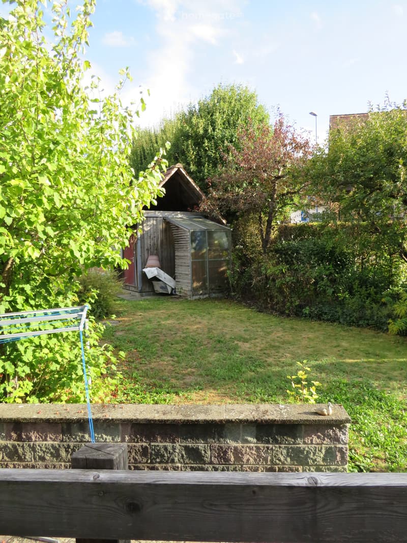 Garden area with a wooden shed, greenhouse, and lush greenery, viewed from a raised platform with a wooden railing.
