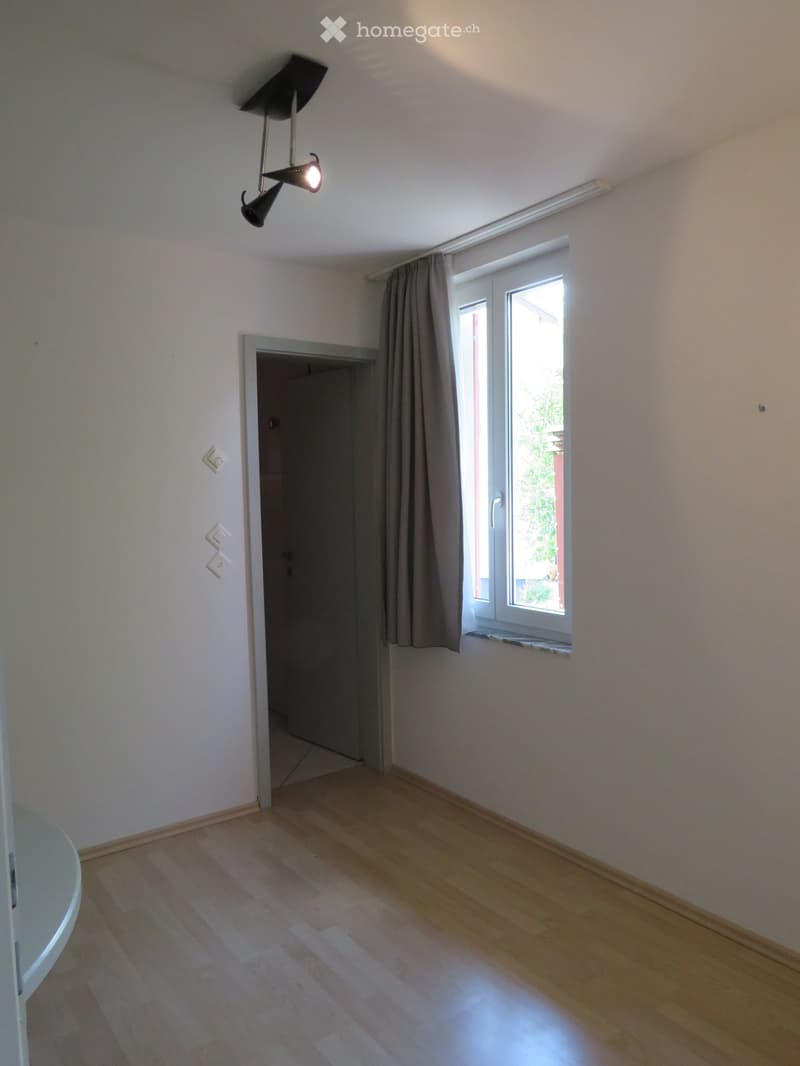 Empty room with wooden flooring, white walls, a window with curtains, and a door leading to another room.