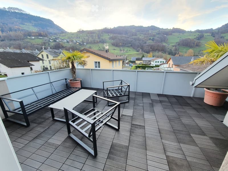 Balcony with wooden decking, modern furniture including a table, chairs, and a bench, with scenic mountain and village views.