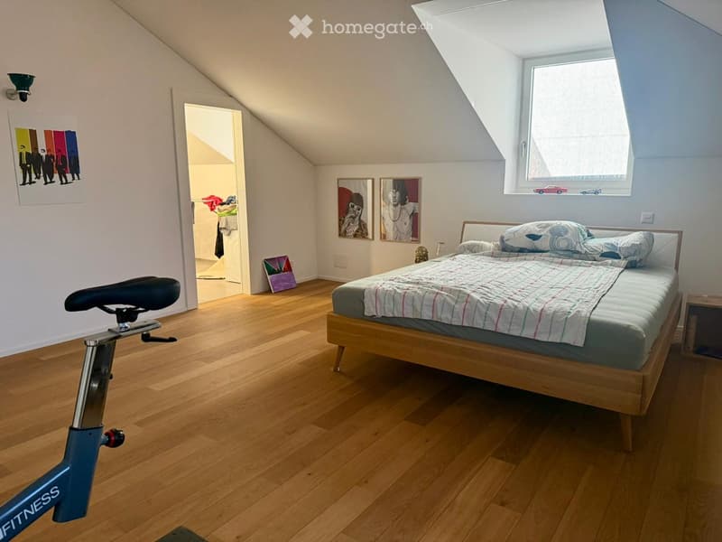 Cozy attic bedroom with wooden flooring, a single bed with a patterned quilt, a nightstand, a bicycle, and several picture frames on the walls.