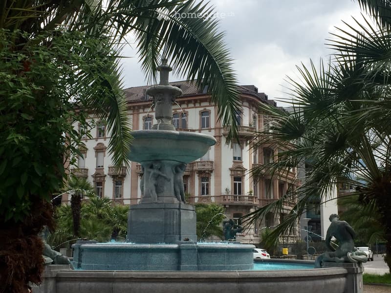 A large stone fountain with multiple tiers and sculptures, surrounded by palm trees and a grand historic building in the background.