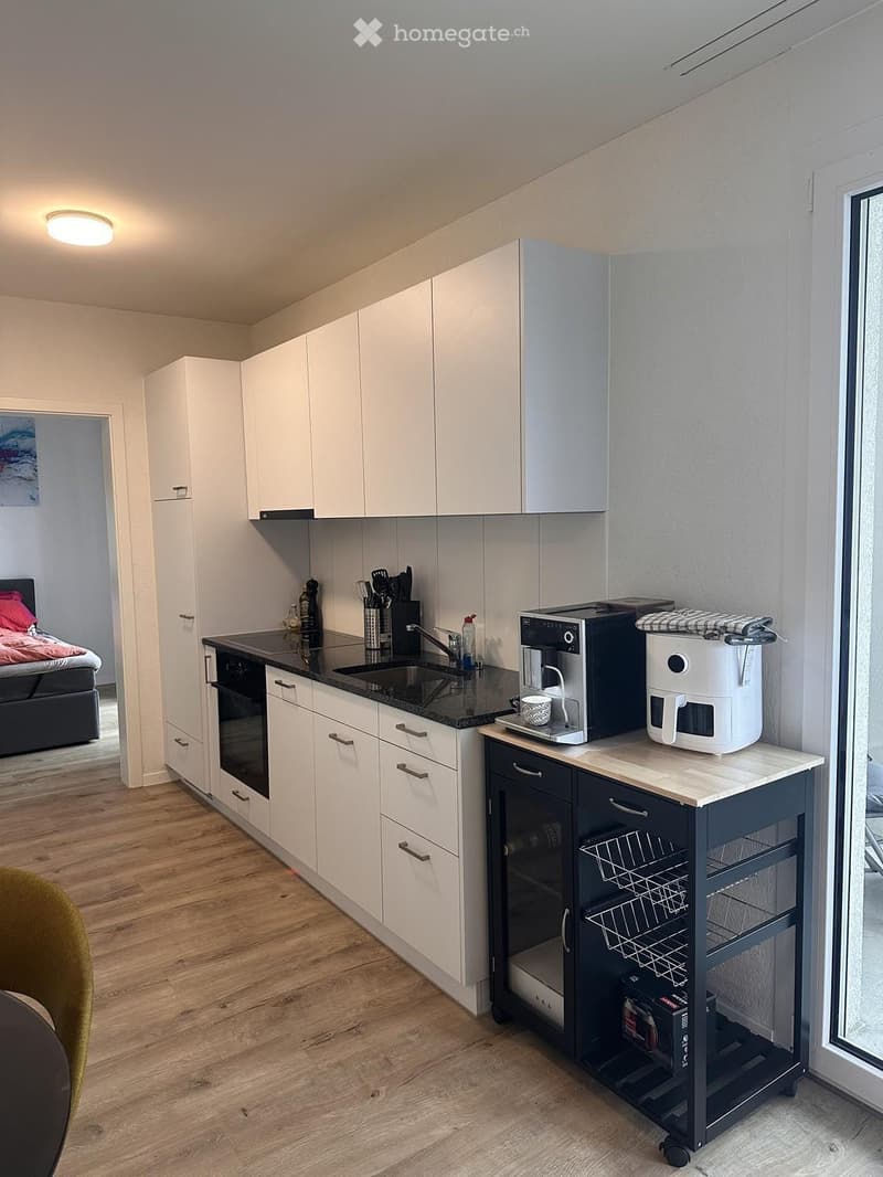 Modern kitchen with white cabinets, black countertop, and stainless steel appliances including a coffee maker and air fryer. A small kitchen cart with wheels is visible, and a glimpse of a bedroom with a bed is seen in the background.