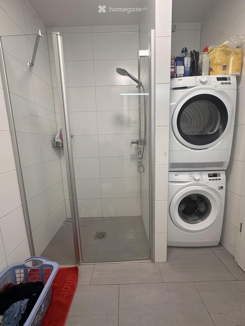 Bathroom with a glass-enclosed shower and a stacked washing machine and dryer unit. Laundry basket with clothes is visible.