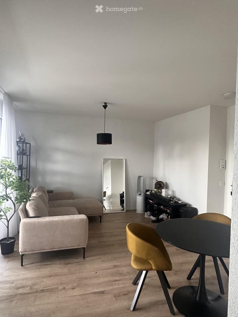 Modern living room with light wooden flooring, a beige sectional sofa, a round black dining table with yellow chairs, a large mirror, a potted plant, and a shelf with decorative items.