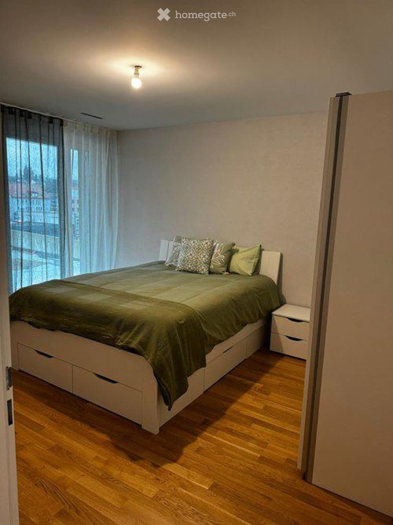 Modern bedroom with a large bed, green bedding, nightstand, and large windows with curtains.