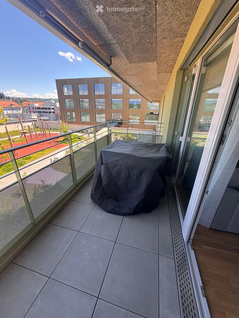Modern balcony with tiled floor, glass railing, covered outdoor furniture, and a view of a contemporary building and park area.