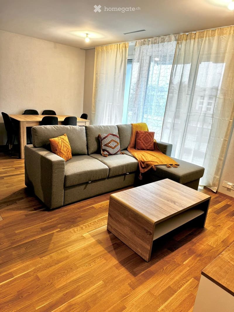 Modern living room with a grey sectional sofa, wooden coffee table, dining table with chairs, and large windows with curtains.