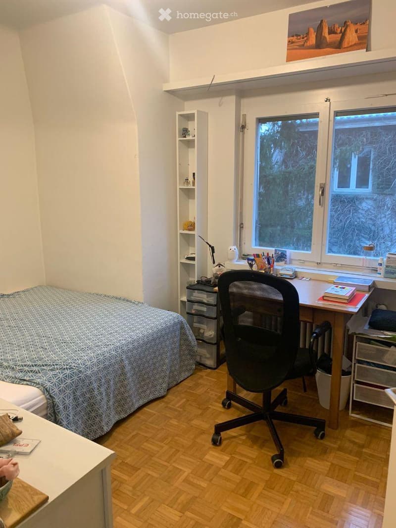 Cozy bedroom with a wooden floor, a bed with a blue patterned duvet, a desk with a chair, a shelf, and a storage unit. Large windows provide natural light.