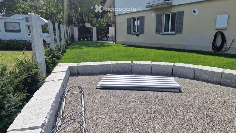 Garden area with a gravel surface, stone border, and a small metal ramp leading to a house entrance. The house has multiple windows and a balcony, with a well-maintained lawn and surrounding greenery.