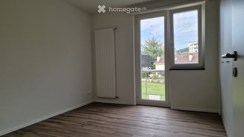 Empty room with wooden flooring, white walls, a radiator, and a large window with a sliding door leading to a balcony.